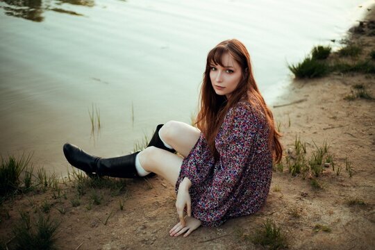 Young woman sitting by a riverbank at sunset, wearing a dress and boots, with water and grass in the background