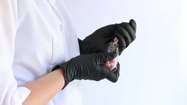 A medical worker holds a transparent jar of pills. A pharmacist in a white coat and black nitrile gloves holds a glass container with pills (health supplements, medicines or vitamins), light backgroun