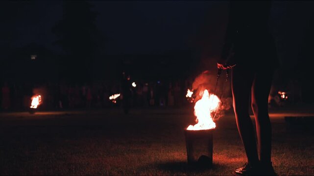 Woman holding flaming torch stands beside a fire pit in a dark outdoor setting, surrounded by a crowd of people enjoying a festive atmosphere