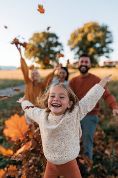 Joyful child in sweater running toward camera while family throws autumn leaves in a golden field on a sunny fall day