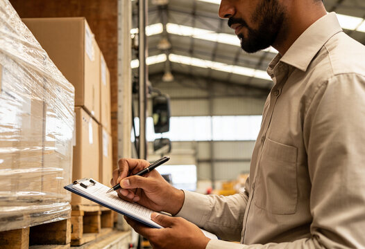 Bearded worker writing on clipboard while checking cardboard box inventory in industrial storage facility warehouse