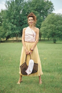 A woman stands in a lush green park, wearing a light pink top and a flowing yellow skirt. She holds a dark tote bag with a white cloth inside, surrounded by trees under a clear blue sky