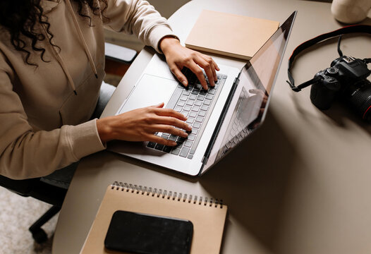 Creative woman typing on laptop keyboard at desk with digital camera and notebook in bright modern home office