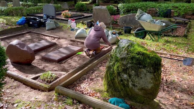 Woman restoring and cleaning a family grave plot in a cemetery, using hand tools beside a wheelbarrow, sand bags, buckets and old stone memorials