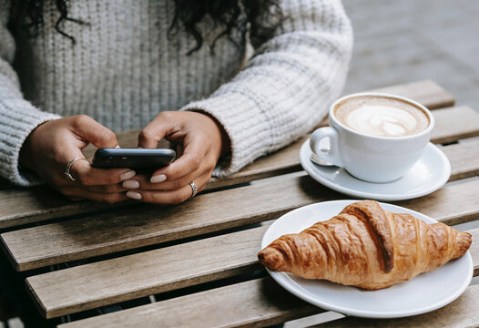 Woman hands using smartphone at outdoor cafe terrace with latte art coffee and fresh butter croissant on wooden table