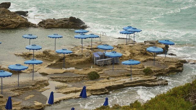 Rocks on the coast with blue parasols and deckchairs, surrounded by the sea with wavy water, Oasis Beach, Kallithea, Rhodes, Dodecanese, Greek Islands, Greece