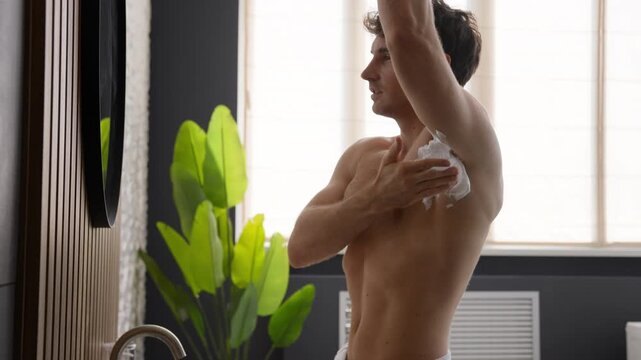 Caucasian millennial man applying shaving gel to armpit in bathroom looking at mirror naked young male guy shawe during morning hygiene routine grooming body care skincare cosmetic procedure at home