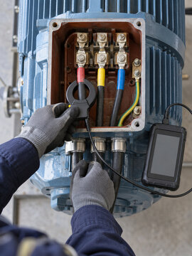A technician tests an industrial motor with a handheld sensor. Gloved hands inspect exposed terminals and electrical cables. Predictive maintenance supports equipment monitoring.