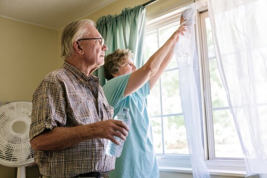 Neighbor helping elderly man stay cool during extreme heat by bringing water and adjusting fan and curtains