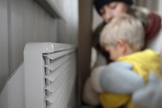 Frozen Mother and Son wearing sweaters shaking and freezing for winter cold embracing over electric heater. Girl and Child is warming hands near Radiator. Discomfort spending time at home. Gas Crisis