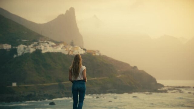 A woman with long blonde hair standing and looking out over the coastal village of Benijo in the Anaga mountain range, Tenerife, Spain, in a cinematic sunset shot with golden haze and misty peaks.