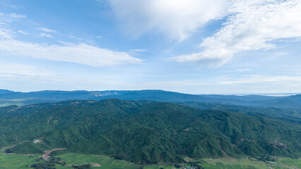 Obraz premium Aerial view of lush green mountain ranges and valleys under a bright blue sky with soft clouds in a scenic landscape
