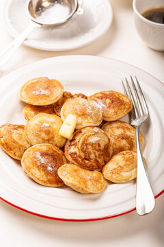 Small Dutch poffertjes pancakes are piled on a white plate with red trim, dusted with powdered sugar and topped with butter, beside a fork, coffee cup, and sugar bowl