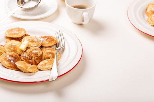 Small golden Dutch poffertjes pancakes sprinkled with powdered sugar fill a white rimmed plate, with butter, fork, coffee cup, with copy space