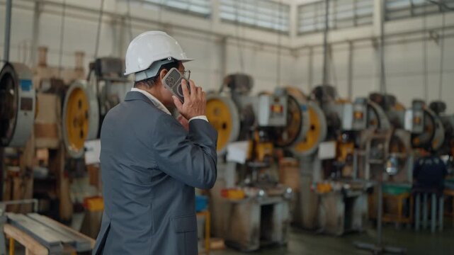 Man wears hard hat, speaks on phone. Large factory looms behind him, industrial. Focus on his actions, not his emotions. Use for business