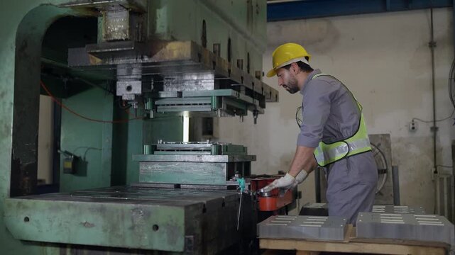 Worker in hard hat and safety vest operates heavy press machine. He adjusts tooling with gloved hands. Factory floor shows worn machinery
