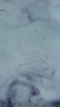 Black ink disperses in clear water, slowly swirling and unfurling into delicate wispy tendrils that spread and intertwine, forming intricate smoke-like patterns against a pale blue backdrop.