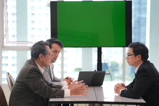 Business meeting with three Asian professionals discussing strategy around conference table in bright office with green screen