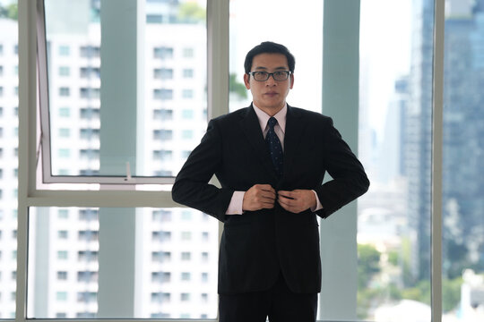 Professional Asian man in black suit buttoning jacket near office window, confident business portrait with urban apartment background and copy space