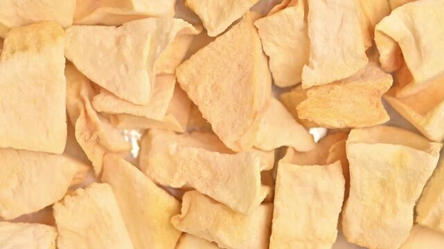 Rotating freeze-dried melon cubes shot in top-down macro close-up under bright studio lighting. Porous yellow dried fruit pieces fill the full frame.
