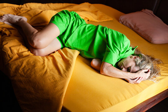 Sad Woman lying curled on a bed with her face covered by her hands. Warm bedding creates an intimate bedroom scene. Visual concept of fatigue, stress, upset or emotional exhaustion.