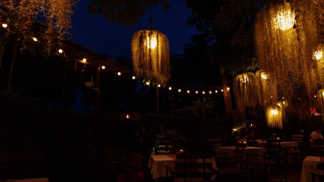 Glowing Hanging Lanterns Illuminate Dining Tables in a Lush Garden Restaurant at Night, Creating a Warm Ambient Setting for Outdoor Dining