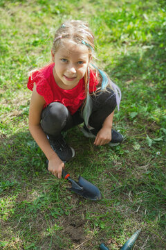 A young girl squats on green grass while holding a gardening trowel during a sunny day. This image represents childhood curiosity, outdoor hobbies, and environmental education for kids.