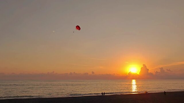 Red parasail gliding across orange evening sky as sun sets over calm ocean, reflecting golden light beside distant beach silhouettes in tranquil coastal landscape