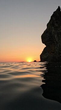 Vibrant sunset glowing over calm sea water beside a towering cliff. Static low-angle shot.