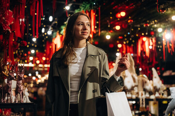 Fototapeta premium Woman carrying shopping bag while browsing brightly decorated festive market with red ornaments, warm lights, and displays in an indoor holiday shopping setting