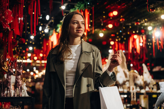 Woman carrying shopping bag while browsing brightly decorated festive market with red ornaments, warm lights, and displays in an indoor holiday shopping setting