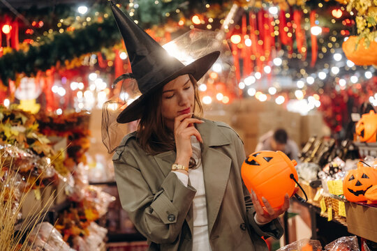 Woman wearing witch hat and holding pumpkin bucket among glowing Halloween decorations in a warmly lit festive retail display
