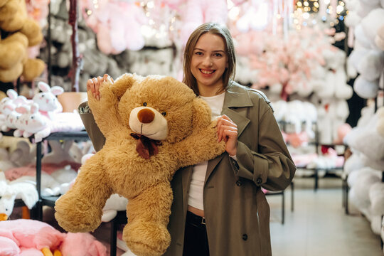 Smiling woman holding oversized brown teddy bear in pastel filled gift shop with plush animals and decorative merchandise, cheerful retail interior scene