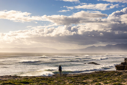 solo female photographer/hiker overlooking cliff with view you massive waves reaching Walker Bay Nature Reserve beach, South Africa at sunset
