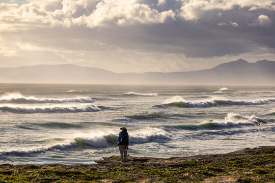 solo female photographer/hiker overlooking cliff with view you massive waves reaching Walker Bay Nature Reserve beach, South Africa at sunset