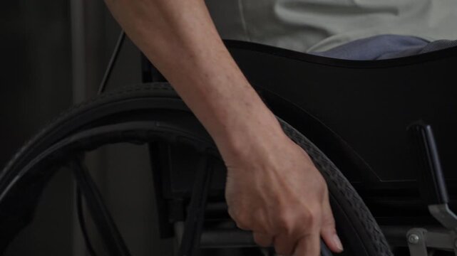 An elderly person sits in a wheelchair alone, hand holding the handrim to move forward slowly, conveying a quiet sense of physical limitation and solitude indoors.