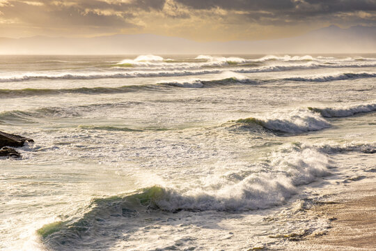 solo female photographer/hiker overlooking cliff with view you massive waves reaching Walker Bay Nature Reserve beach, South Africa at sunset