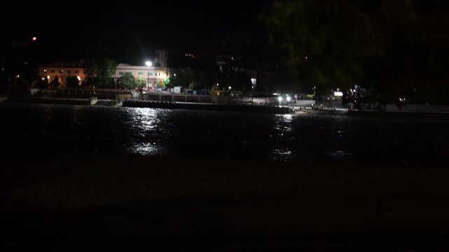 Overhanging foliage frames bright lights along the opposite bank of the Ganga River in Rishikesh at night.