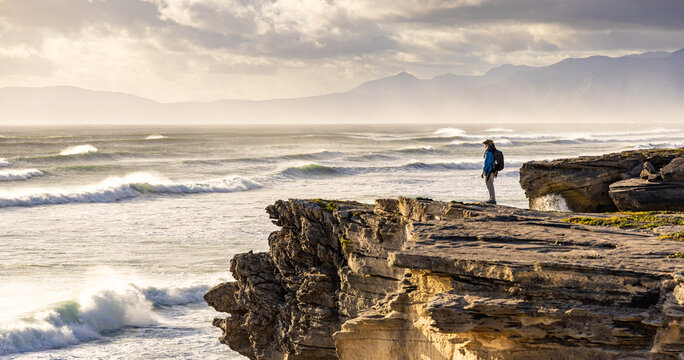 solo female photographer/hiker overlooking cliff with view you massive waves reaching Walker Bay Nature Reserve beach, South Africa at sunset