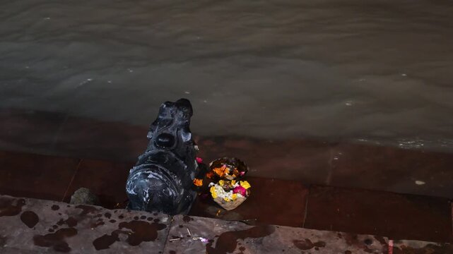 A black Nandi bull sculpture stands beside flower offerings at the edge of the Ganga River in Rishikesh at night.