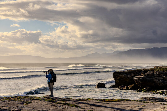solo female photographer/hiker overlooking cliff with view you massive waves reaching Walker Bay Nature Reserve beach, South Africa at sunset