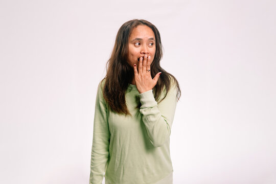 Portrait of a surprised Asian woman covering her mouth with one hand and looking to the side on a white backdrop.
