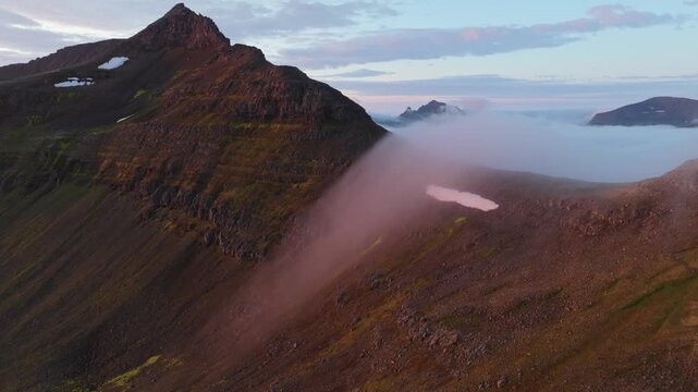 Aerial view boom up, pull back of rugged Hornstrandir, Iceland, with a dramatic cloud inversion filling the mountain pass at sunset. A remote, peaceful, and stunning landscape.