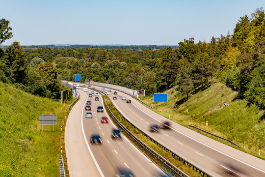 High angle view of multi lane highway winding through green forest with motion blur cars under sunny blue sky, German autobahn and road transport concept