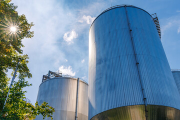 Low angle view of large metallic industrial storage silos and tanks with bright sunburst through green tree leaves against blue sky, modern industrial facility and renewable energy concept © JaRu Creative Studio