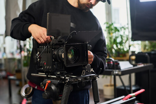 Young adult Caucasian male videographer adjusting professional cinema camera equipment on set. Useful for film production, broadcasting services, digital cinematography marketing