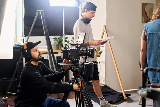 Caucasian young adult videographer operating professional cinema camera on tripod during film production with crew. Useful for media advertising