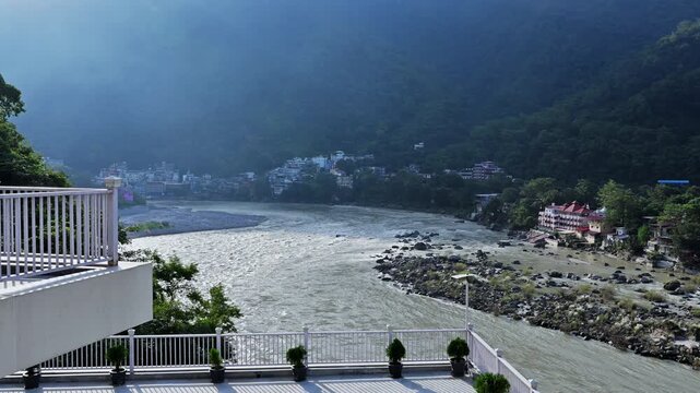 Sunlit haze hangs above the Ganga River and wooded slopes beyond white riverside terraces in Rishikesh.