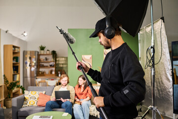 Young adult Caucasian sound technician holding boom microphone while filming young adult actors indoors. Suitable for video production, broadcasting, media industry advertising © pressmaster