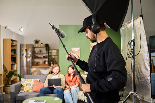 Young adult Caucasian sound technician holding boom microphone while filming young adult actors indoors. Suitable for video production, broadcasting, media industry advertising
